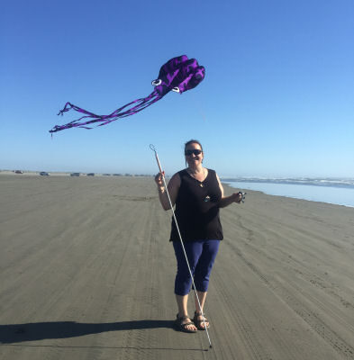 Gina, standing on the beach, smiling, holding her cane in her right hand, while her left hand holds the string to a kit flying high behind her - a purple Octopus she calls Mr. Squiggly