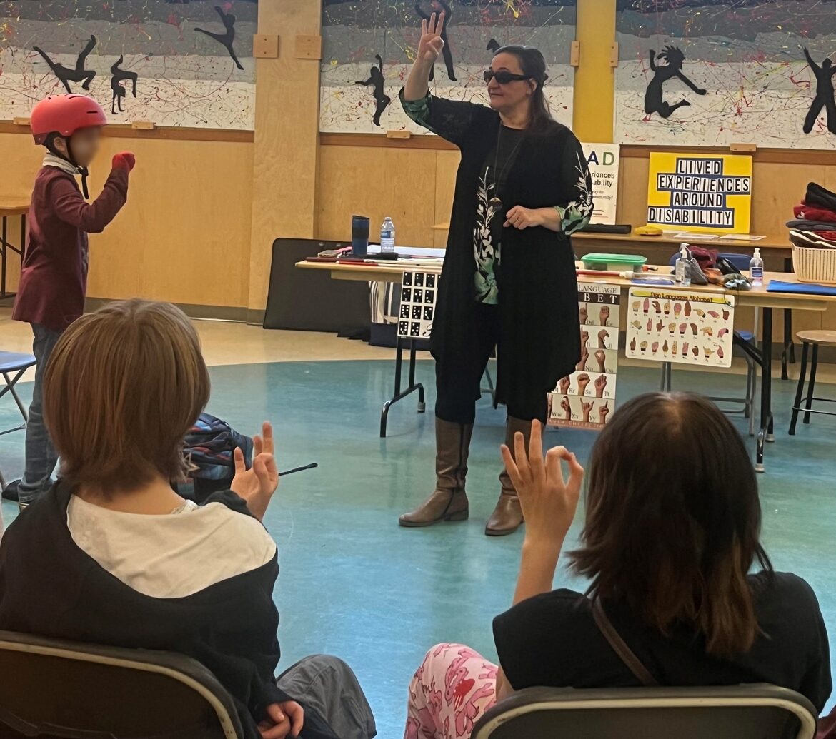 Gina standing at front of a room filled with students during a school presentation. She is holding up her hand, teaching the students how to form the letter F in sign language, using their hands.