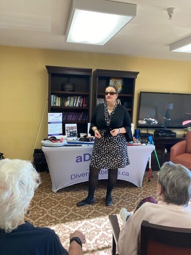 Gina is standing in front of her props table and presenting in the library at a seniors retirement community. She is wearing a black and white dress with a short cropped black top.