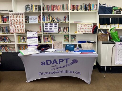 Youth adapt table display. Props and material are displayed on a table with a covering saying.aDAPT DiverseAbilities.ca. There is a sign language and braille poster behind the table that is part of the program teaching. 