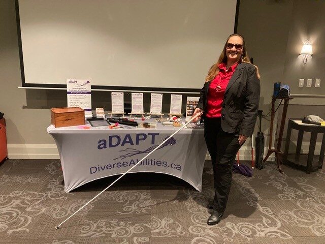 Gina standing in-front of her table lined with props for her Vision loss presentation. She is holding her white cane, wearing her dark glasses and smiling. 